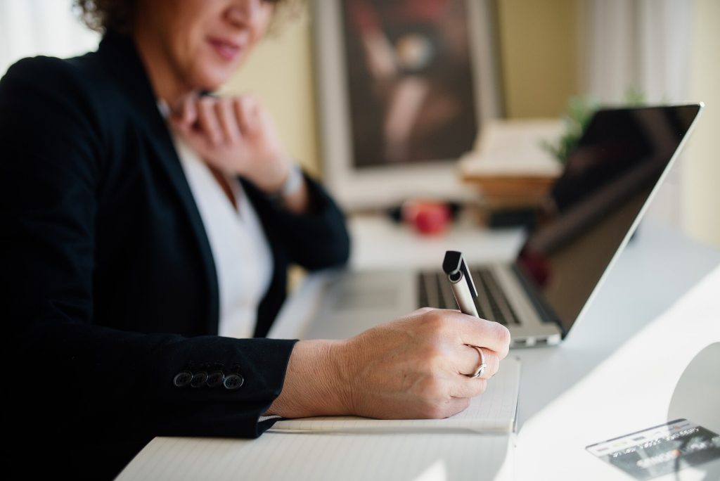 A business professional is seated at a sleek desk, actively taking notes with a pen while focused on a laptop screen. Natural light enhances the workspace's ambiance.