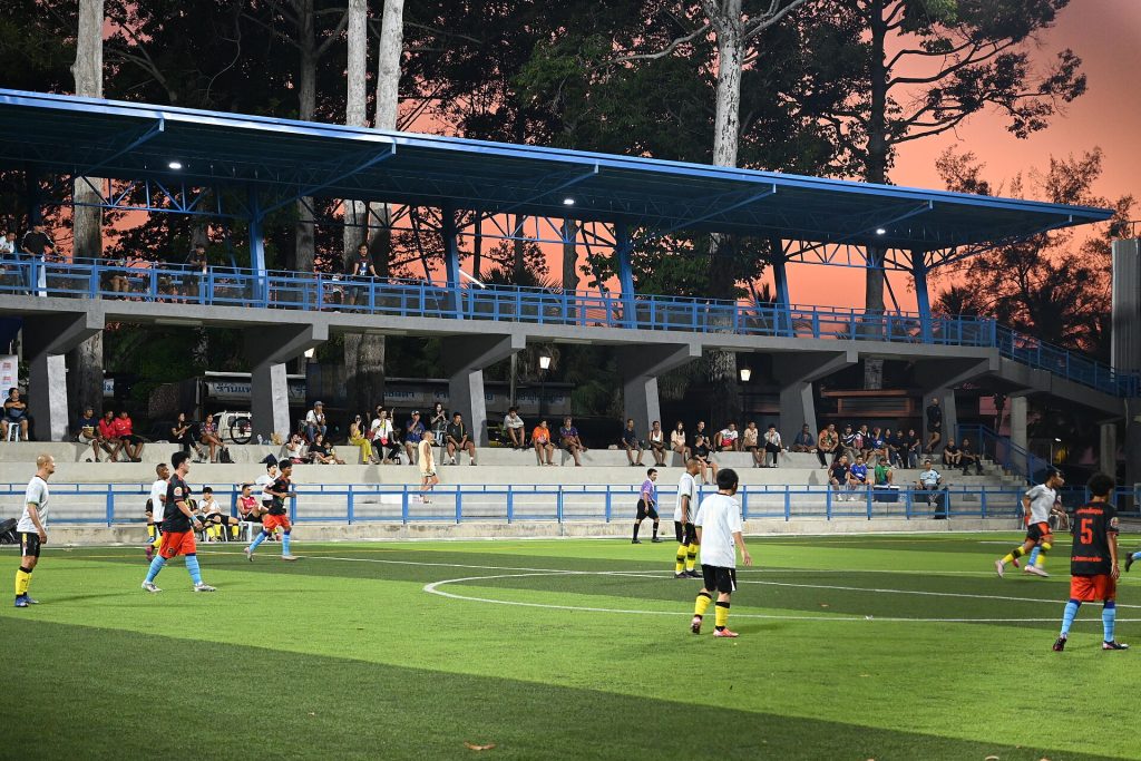 This photo shows a football (soccer) match being played at a local stadium during what appears to be late afternoon or early evening, given the warm, golden lighting. The scene captures players on a well-maintained grass pitch, with some wearing different colored jerseys suggesting this is an active game in progress. The stadium features a modest but functional design with a covered grandstand that has a distinctive blue roof structure supported by concrete pillars. Spectators can be seen seated in the stands watching the match. The facility appears to be a community-level sports venue rather than a major professional stadium. The lighting creates a pleasant atmosphere with warm tones visible in the sky and on the surrounding buildings in the background. Trees are visible beyond the stadium structure, and the overall setting suggests this is located in a residential or mixed-use area. The photo captures the essence of grassroots football culture, where local communities gather to enjoy the sport in a more intimate setting than large commercial stadiums.