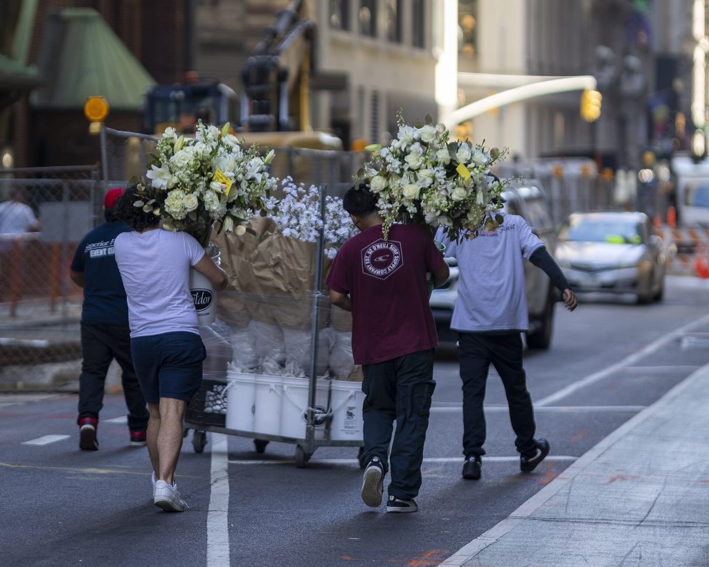 Workers push a rolling cart of large white floral arrangements down a street in New York City.