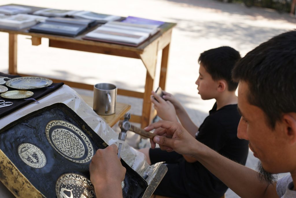 An artisan works by hand on a decorative metal object, engraving or chasing its surface with small tools. The scene documents precision metalwork, workshop practice and the continuation of applied-art traditions in Bukhara.