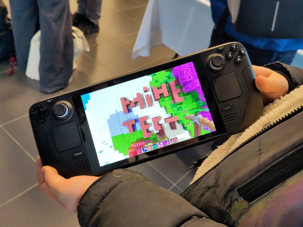 Minetest on a Steam Deck device, shown during the FOSDEM conference about Free and Open Source software in 2024. The person is holding the device with their hands. They are in front of the official Minetest booth at FOSDEM 2024. Photo taken from a Fairphone.
