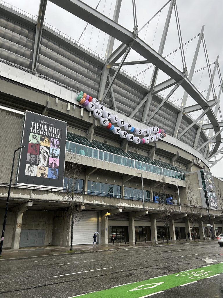 A friendship bracelet reading "TAYLOR SWIFT THE ERAS TOUR" on the side of BC Place, along with a poster for the tour.
