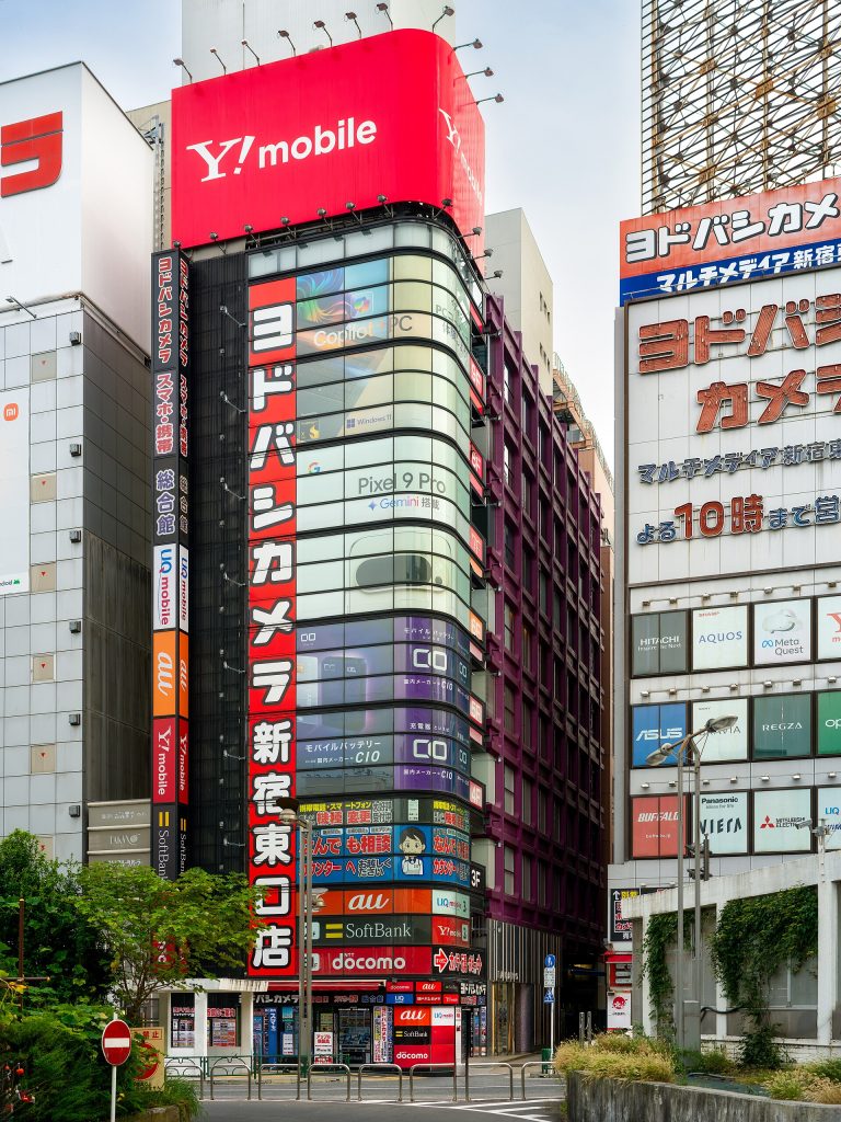 Located in the heart of Tokyo’s vibrant Shinjuku district, this scene captures the iconic Yodobashi Camera store, a multistory haven for technology enthusiasts and one of the city’s must-visit destinations for electronics. Its sleek modern facade, adorned with bold red signage and floor-to-ceiling glass panels, reflects Tokyo’s high-tech culture and fast-paced energy. This building stands as a symbol of Japan’s technological prowess, offering everything from the latest smartphones to high-end cameras and gaming equipment. Yodobashi Camera’s architectural design exemplifies functional modernism. The transparent glass facade invites passersby to peer into the buzzing retail spaces within, creating an atmosphere of accessibility and excitement. The vibrant advertisements and colorful logos covering the building are quintessential Tokyo, contributing to the city’s reputation as a visual feast of neon and bold typography. The surrounding area, bustling with other electronic stores and department buildings, highlights Shinjuku’s status as a commercial powerhouse. Historically, Shinjuku evolved as a major transportation and business hub, and today it represents a seamless blend of tradition and innovation. The juxtaposition of lush greenery at the street level with towering steel-and-glass structures emphasizes Tokyo’s architectural duality, where nature and urban development coexist. Visiting this area offers more than just shopping—it’s an opportunity to immerse yourself in Tokyo’s dynamic cityscape, where architecture, commerce, and culture intersect. Whether you’re looking to explore cutting-edge technology or simply soak in the energy of one of Tokyo’s busiest districts, Yodobashi Camera and its surroundings are an unmissable part of the Shinjuku experience.