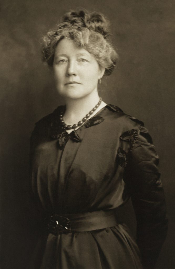 Ada S. Flatman by Harris & Ewing in a 3.5 x 5 in photographic print. A cropped version of this photograph was published in The Suffragist 5, no. 58 (Mar. 3, 1917): 10. The caption on an enlarged, cropped version of the identical image in the same folder reads: "Miss S. Ada Flatman, of London, England, is Advertising and Business Manager of 'The Suffragist,' official weekly organ of the Congressional Union for Woman Suffrage and the National Woman's Party." Formal portrait, half-length, standing with arms pulled behind back, wearing dark, belted dress with stain trim, small bow at v-neck, and beaded necklace.