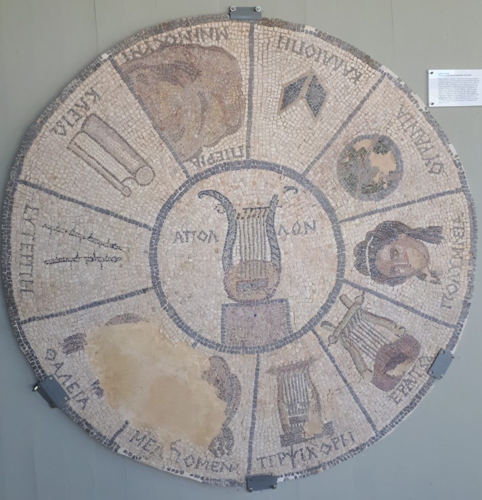 Mosaic disc with the symbols of the nine Muses and their mother Mnemosyne: Calliope (writing board), Urania (globe), Polyhymnia (pensive face), Erato (lyre), Terpsichore (cithara), Melpomene (tragic mask), Thalia (comic mask), Euterpe (flutes), Clio (scroll), and Mnemosyne (Mt. Pieria). Apollo's lyre in the middle. Archaeological Museum of Elis.